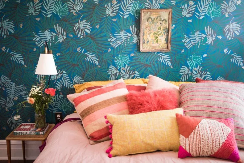 Bedroom with blue-and-green leafy wallpaper, pink bedding, yellow and striped cushions, wooden bedside table and white-shaded lamp.