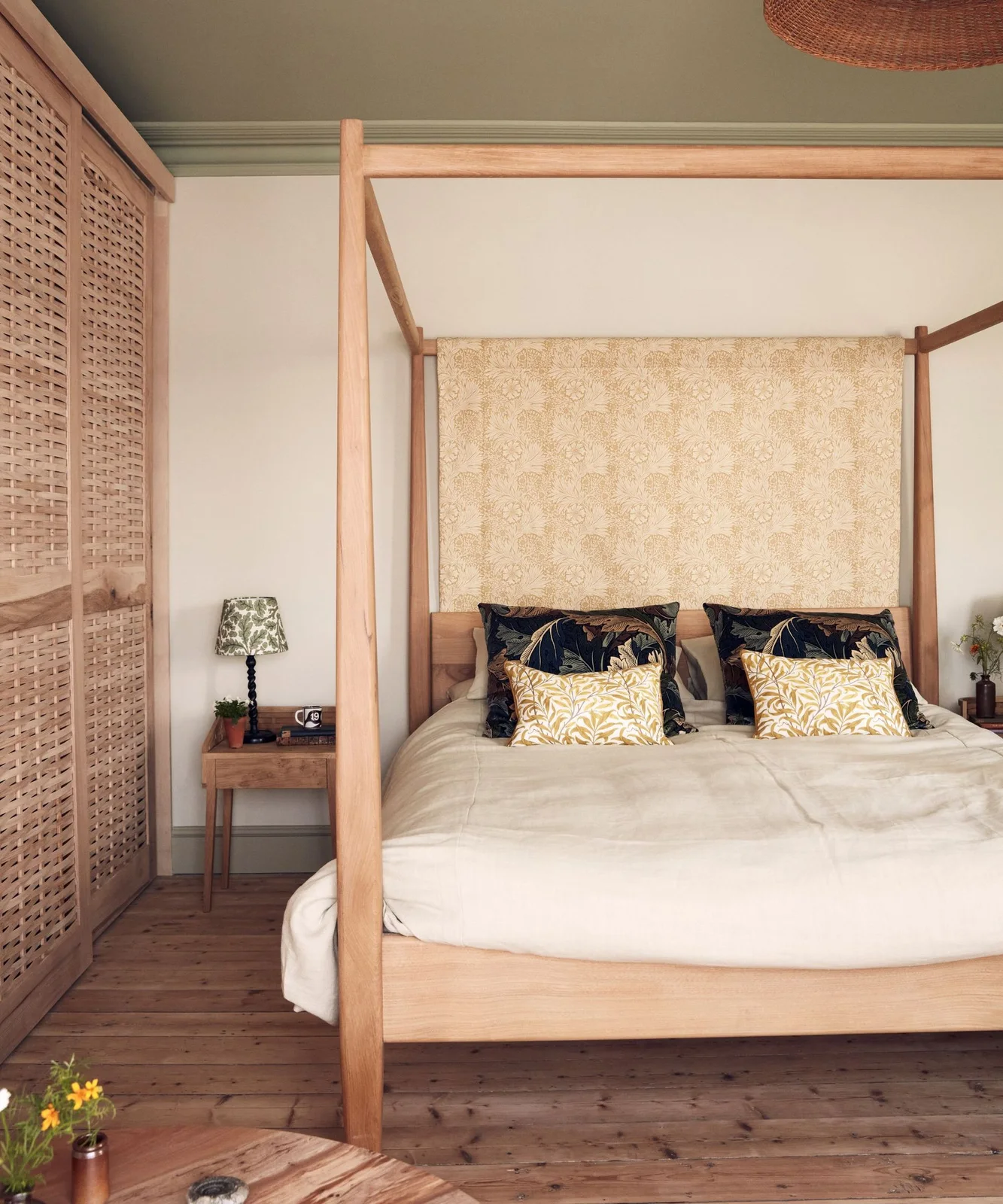 A wooden canopy bed with pale bedding sits beside woven-fronted storage beneath a pale patterned fabric backdrop and darker botanical cushions