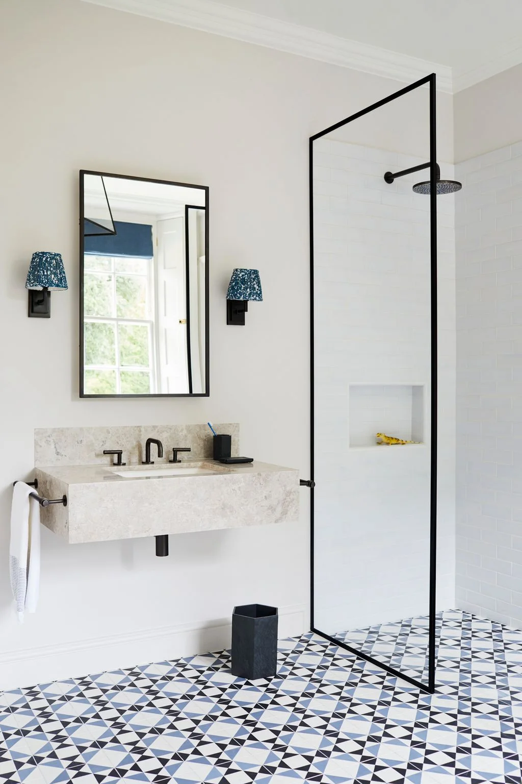A narrow black-framed fixed screen stands between a floating sink and a white tiled shower above a blue and black patterned floor.
