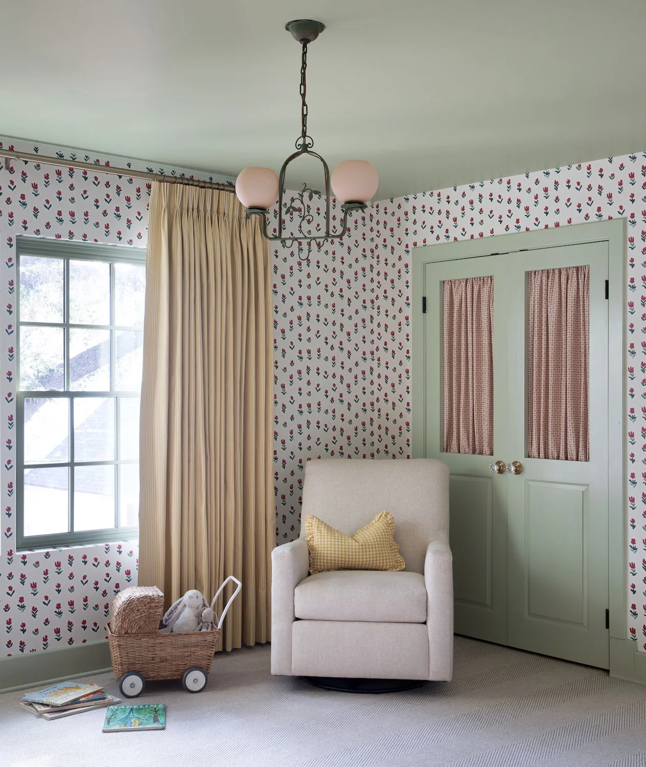 Small red floral wallpaper surrounds pale green doors and ceiling beside yellow curtains, a pale adult chair and pink globe lights