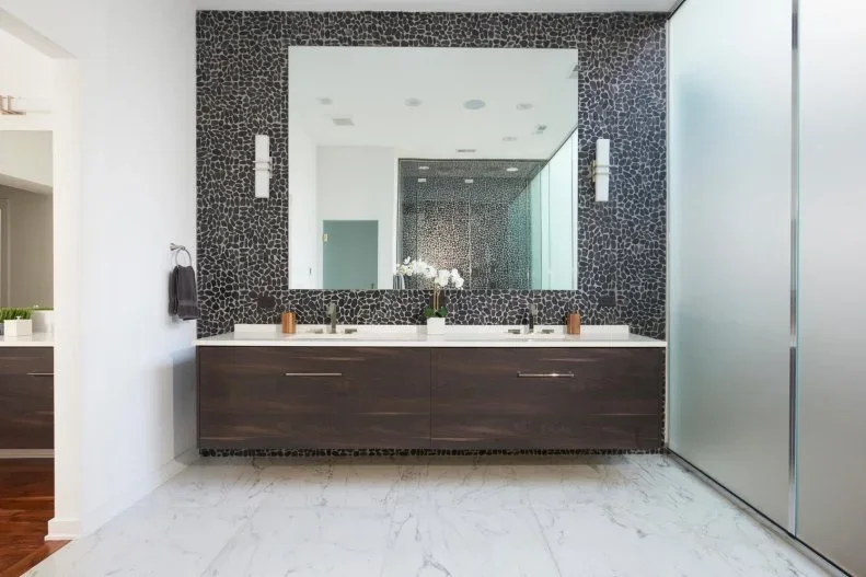 A dark floating bathroom vanity with two sinks sits beneath a large mirror on a dark small-scale textured wall beside pale veined flooring and glass shower panels
