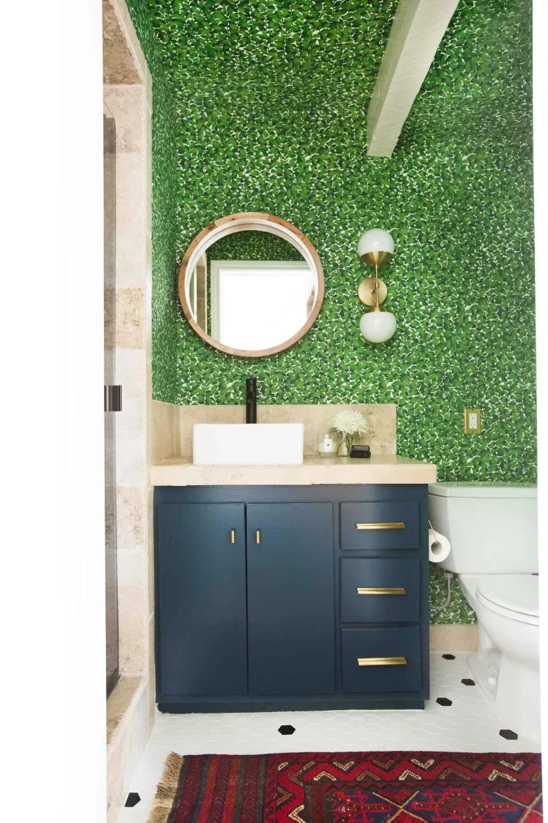 Dense green leaf wallpaper covers walls and ceiling above a navy vanity, round mirror and white floor with small dark accents, with a red patterned rug below.