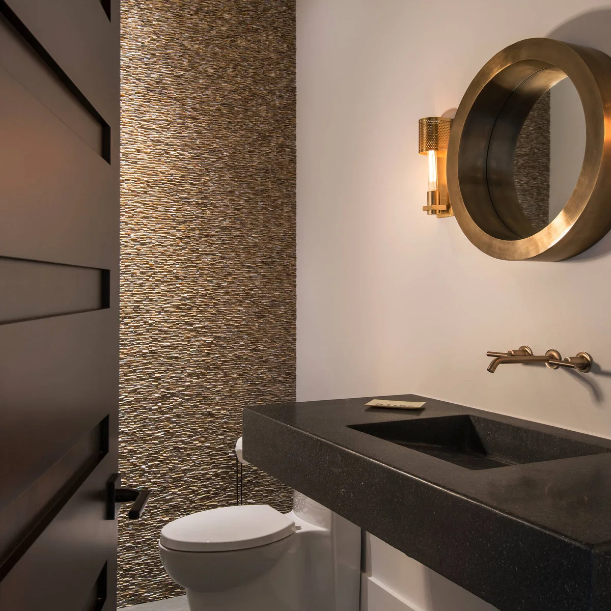 A thick dark wall-mounted basin beneath a round warm-metal mirror, beside a textured wall and wall-hung toilet.