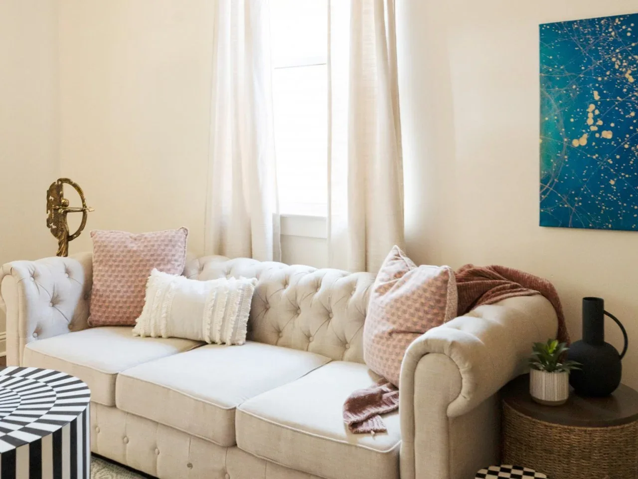 A pale buttoned sofa with pink cushions sits between a woven side table and a black-and-white patterned coffee table.