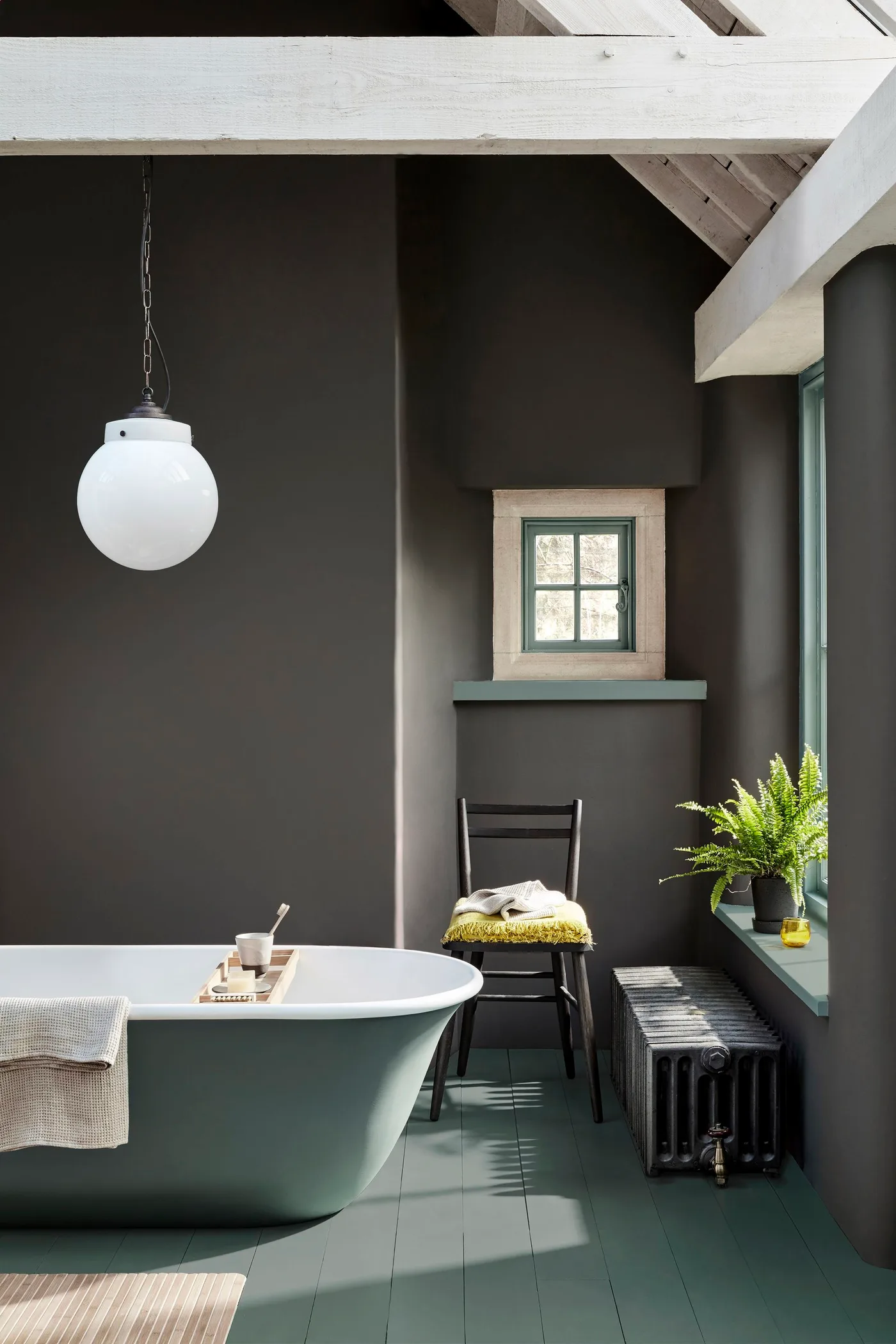 Bathroom with deep warm gray walls, green-gray freestanding bath and floor, white ceiling beams, globe pendant, black chair, and fern.