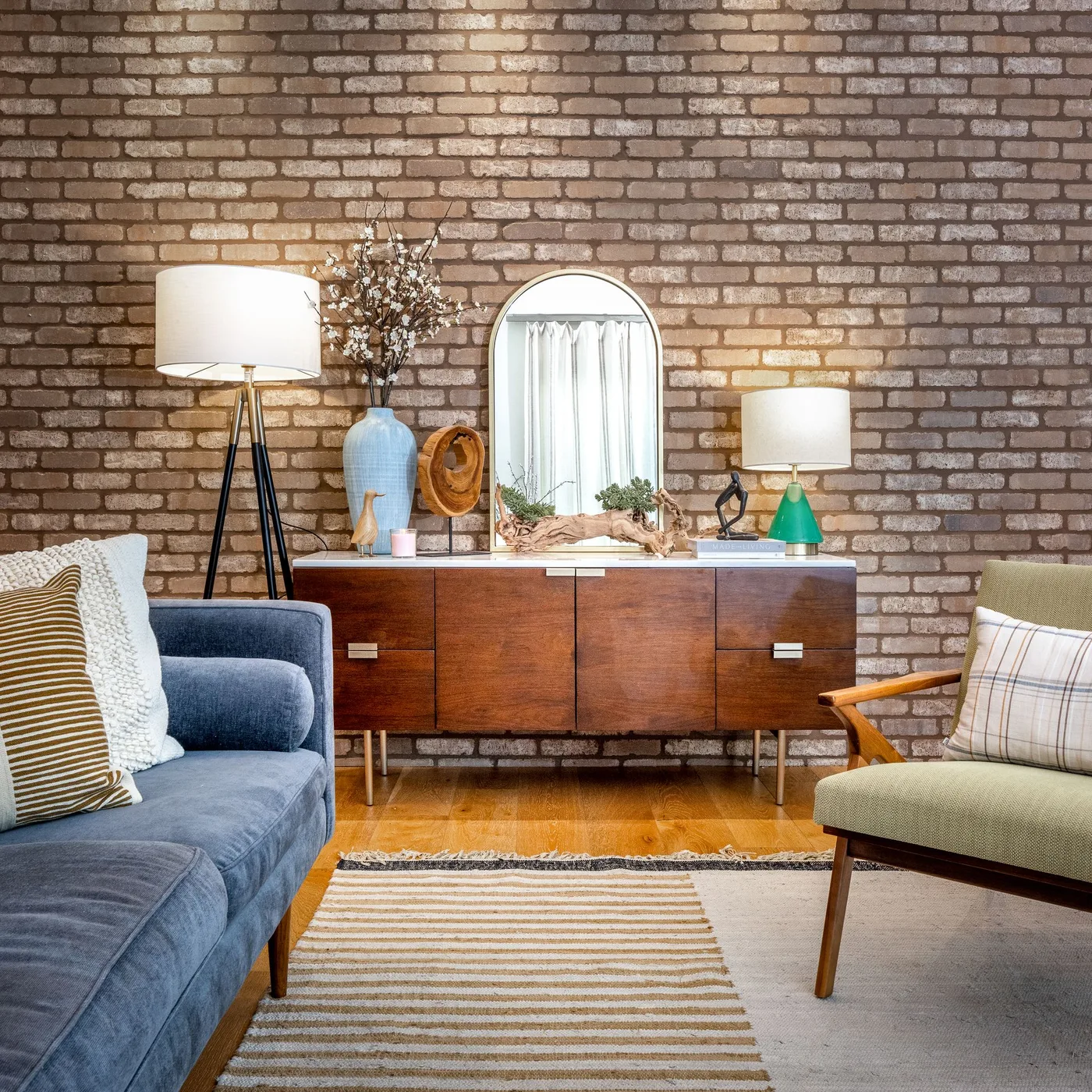 Warm brick living room wall behind a wood sideboard, arched mirror, blue sofa and pale green chair