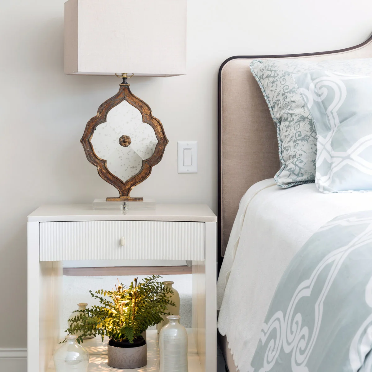 White bedside table with ribbed drawer front beneath a decorative mirrored lamp