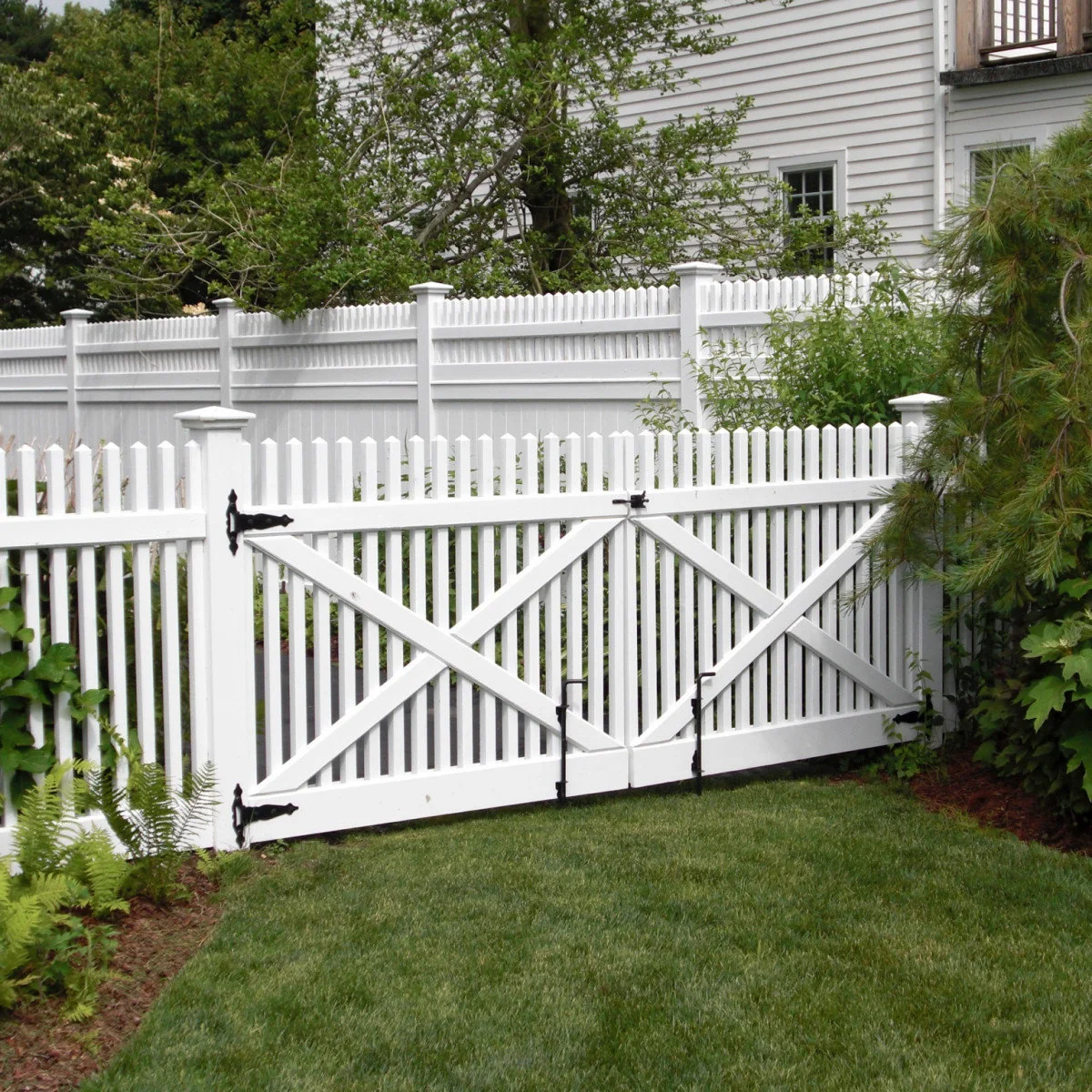 A pair of white picket gates with diagonal braces and dark hinges in a planted front garden.