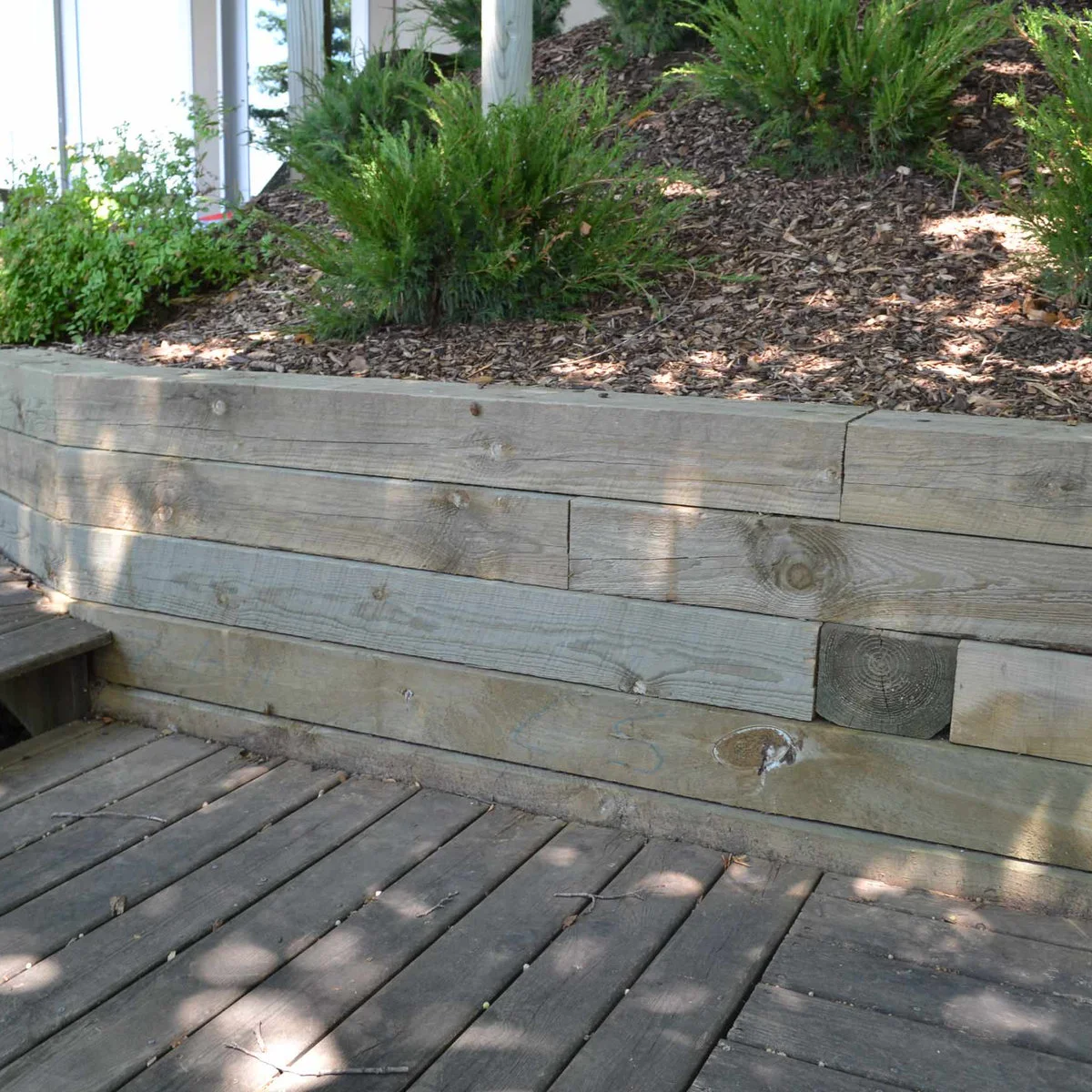 Horizontal timber courses retain a mulched planting bed beside a wood deck.