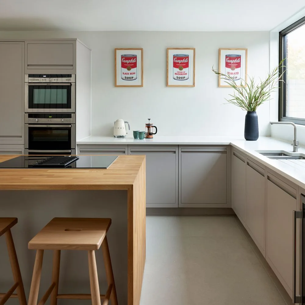 Flat pale gray cabinets and recessed pulls surround built-in ovens beside a wooden-topped island beneath three framed prints