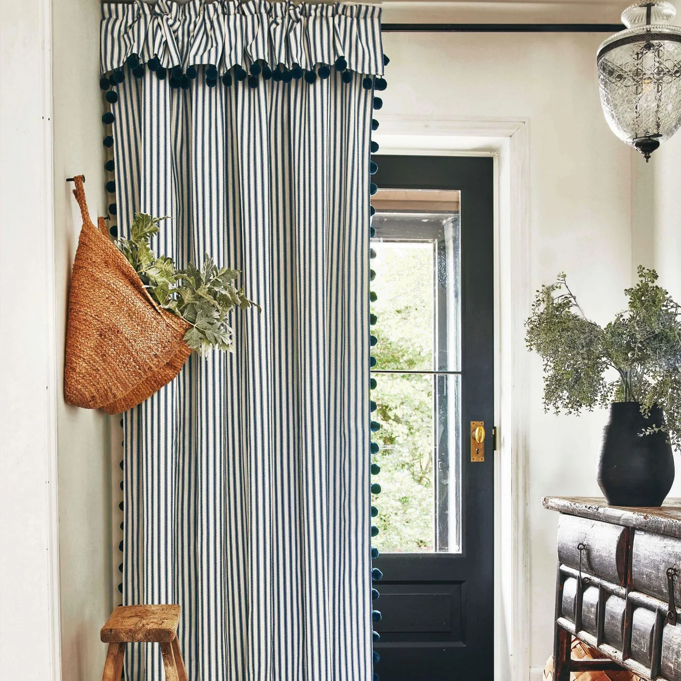 Entrance with a blue striped curtain and dark trim, black glazed door, wooden console, small wooden stool and hanging woven bag.