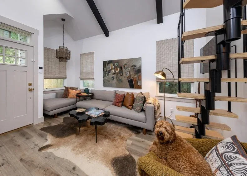 A pale sectional sofa and small dark tables sit below simple wall artwork beside an open staircase, wood floor and pale window shades