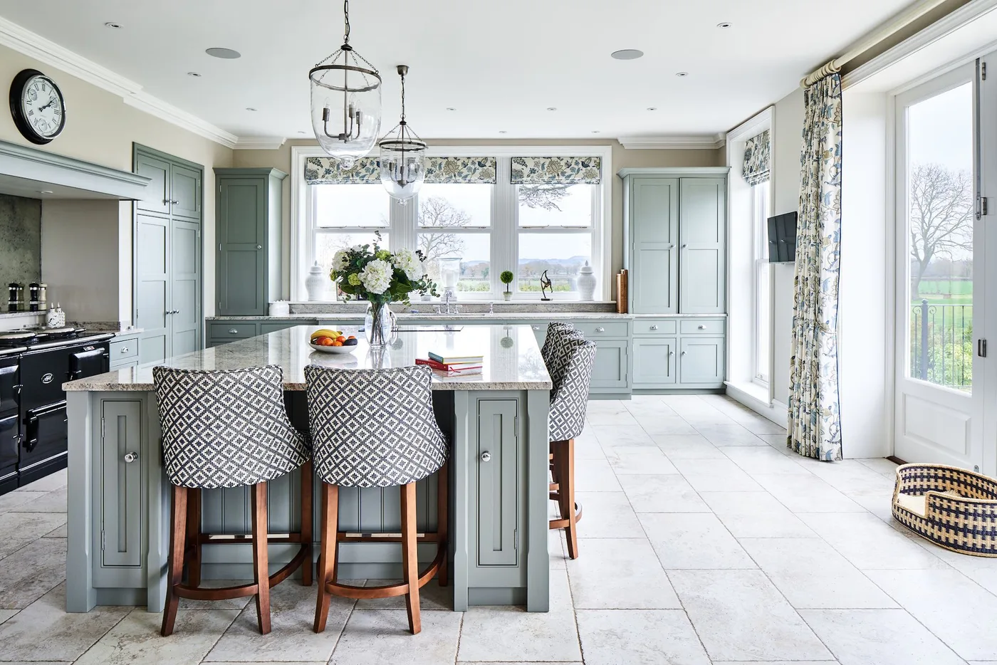 Pale green kitchen with island, patterned upholstered stools, light floor and floral window treatments.