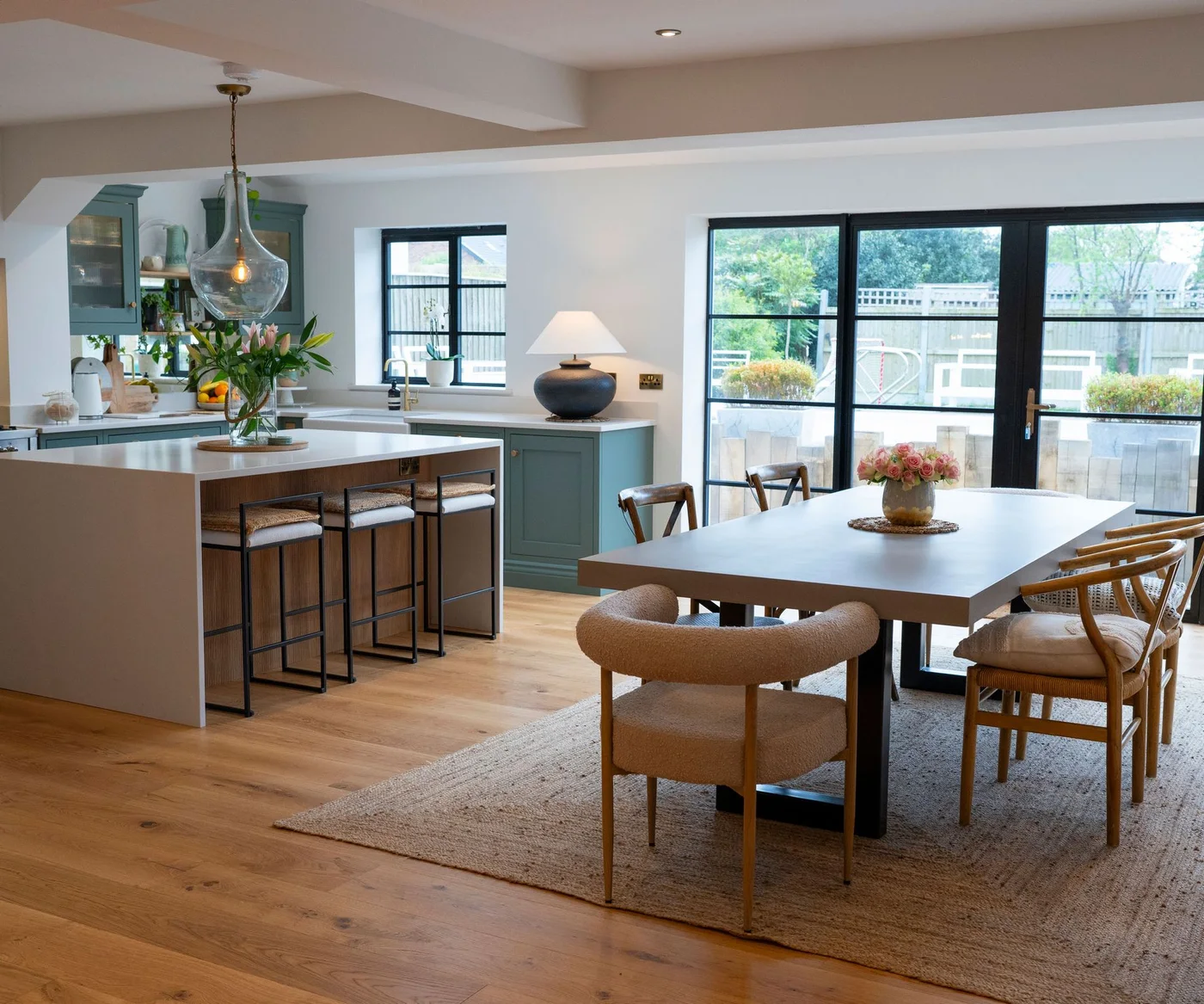 A rectangular dining table stands on a pale rug beside a wooden island, green kitchen cabinets and black-framed garden doors
