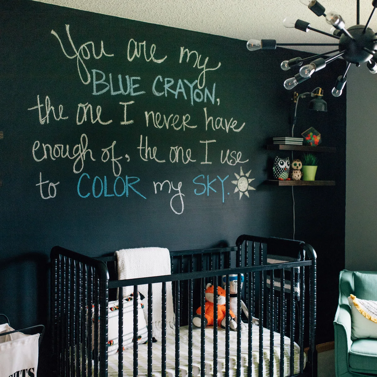Black crib against a dark green wall with pale writing, a mint-colored chair and a black ceiling light