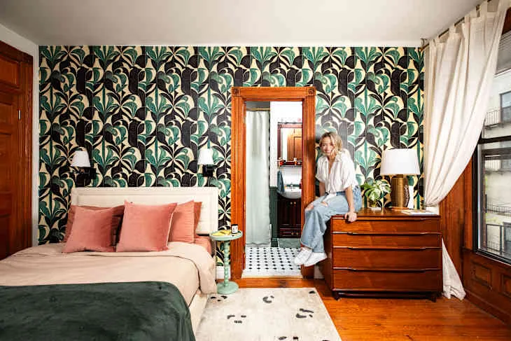 Bedroom with dark green botanical wallpaper, pale bed, pink cushions, green throw, wooden dresser and broad wooden mirror frame.