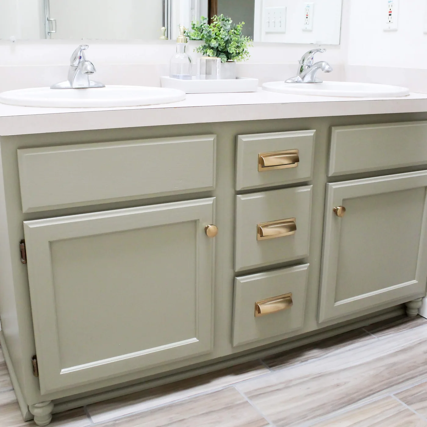 Pale green painted double vanity with gold-toned cup pulls and knobs beneath an existing white counter and two basins.