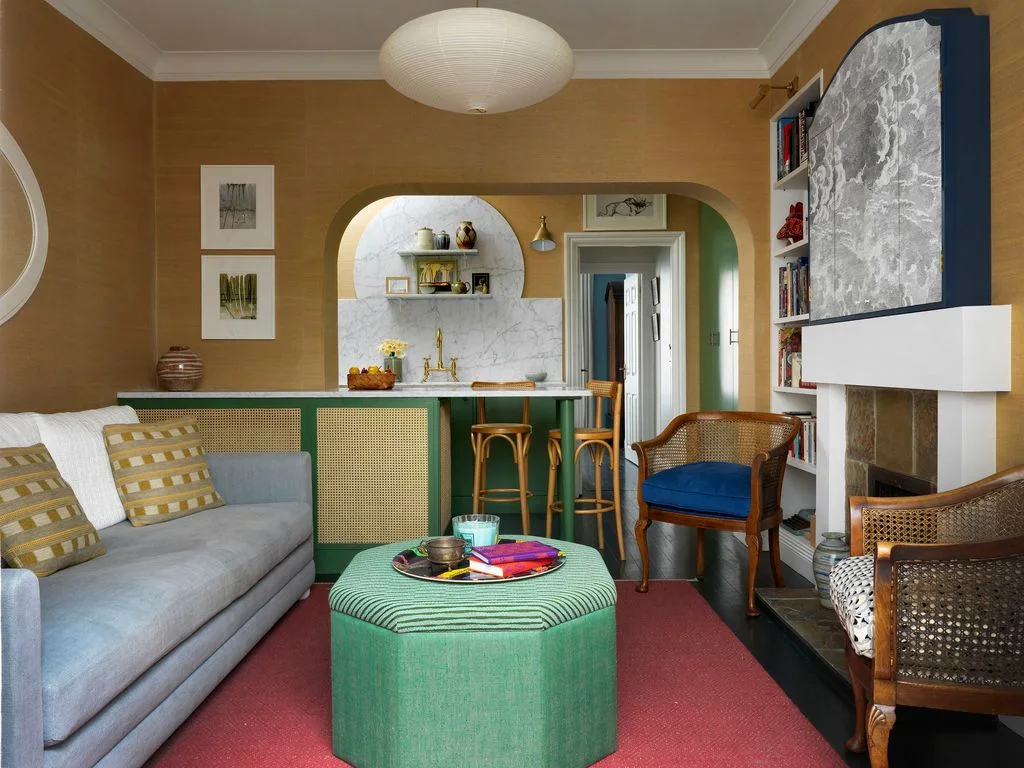 A green peninsula with woven front panels separates a small kitchen from a gray sofa, green ottoman and timber chairs.