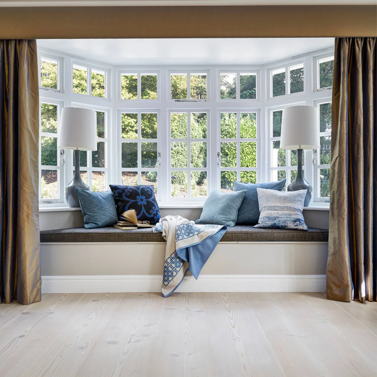 A gray window-seat cushion with blue pillows, two lamps and long curtains framing a many-paned bay.