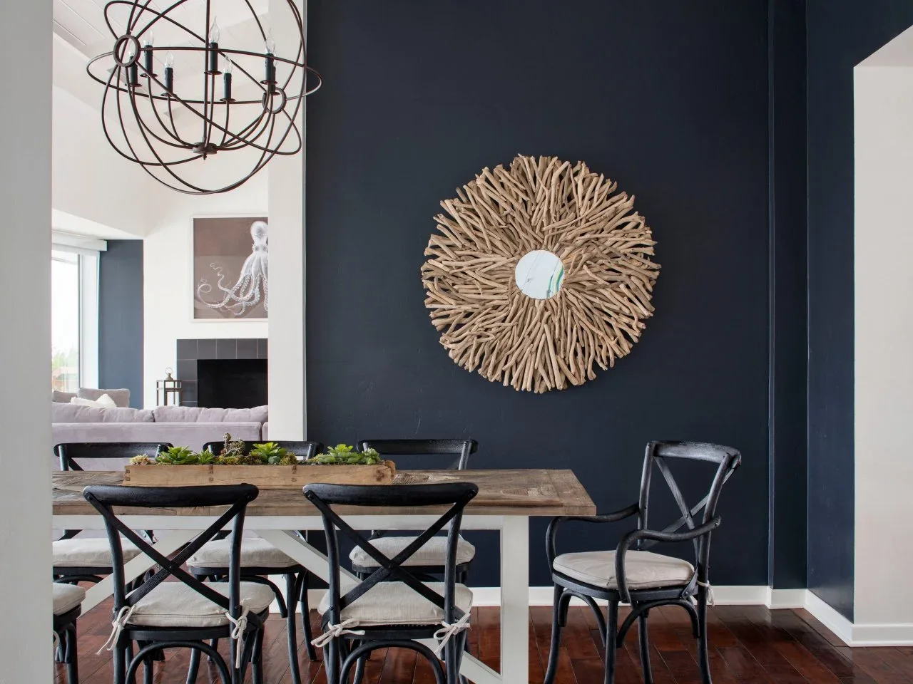 A navy dining wall holds a broad twig-like round mirror above a timber table with dark cross-back chairs and pale seat pads.