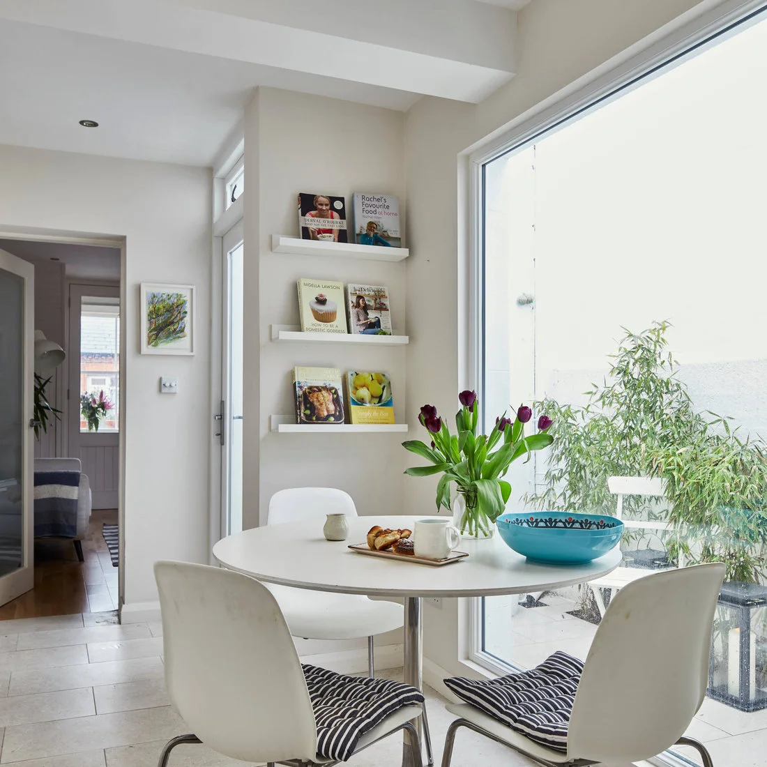 A round white table and white chairs sit beside a broad window and narrow cookbook display ledges.