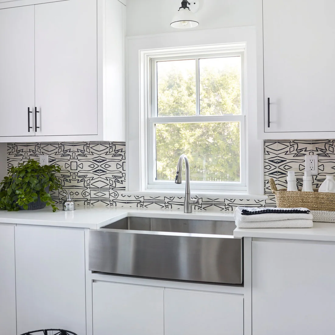 A deep metal utility sink with a tall tap sits beneath a window between white cabinets with dark pulls and a patterned backsplash.