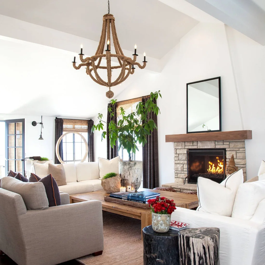 Pale living-room sofas and a gray chair surround a wooden coffee table beneath a rope-like chandelier beside woven blinds and a textured fireplace