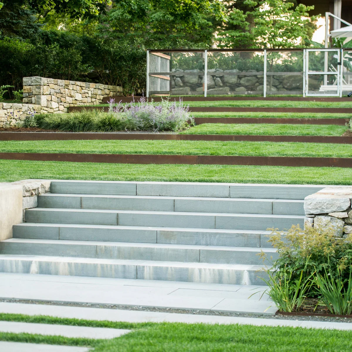 Narrow dark retaining edges form stepped lawn terraces behind broad pale steps and beside a stone wall.