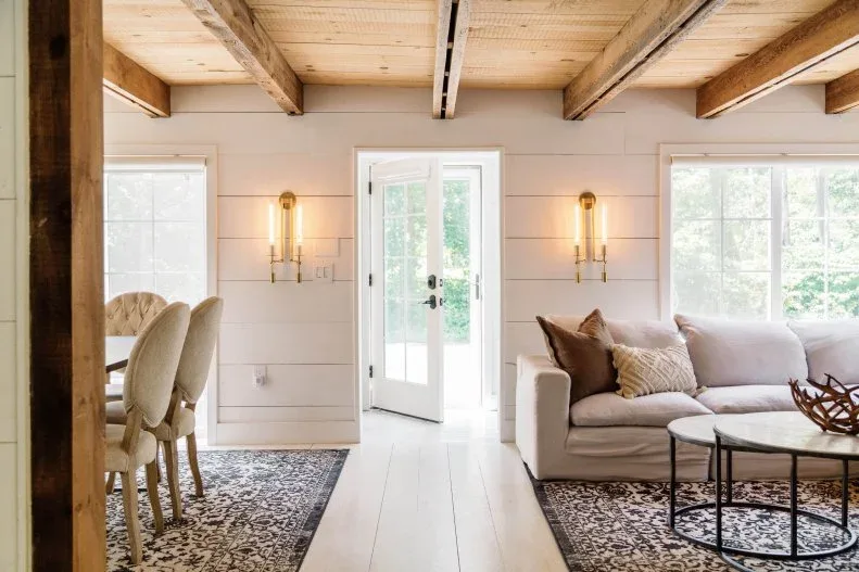 A glazed entrance door between white paneled walls and living room seating beneath timber beams