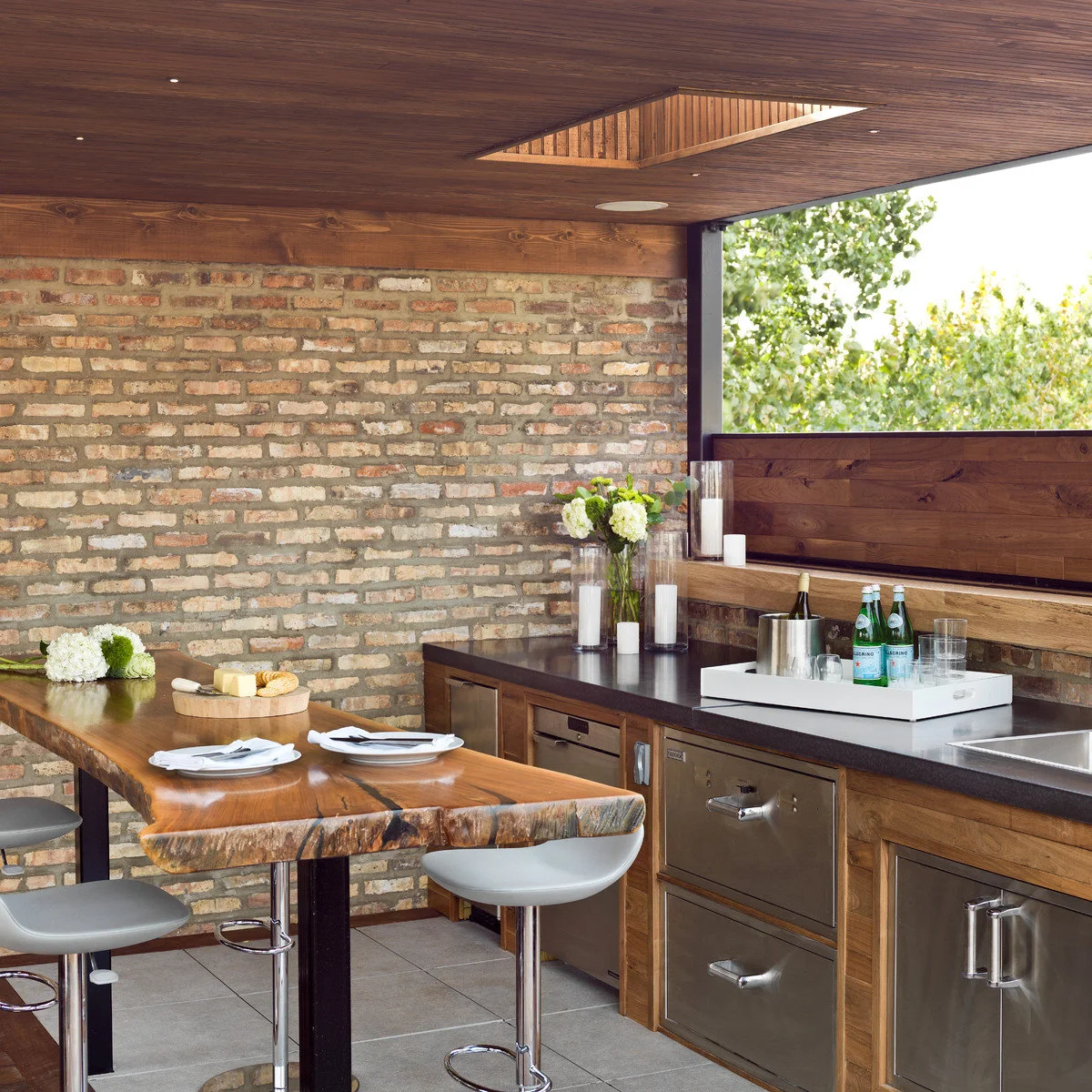 Sheltered outdoor preparation area with brick wall, wood cabinets, dark counter, sink and thick wood table