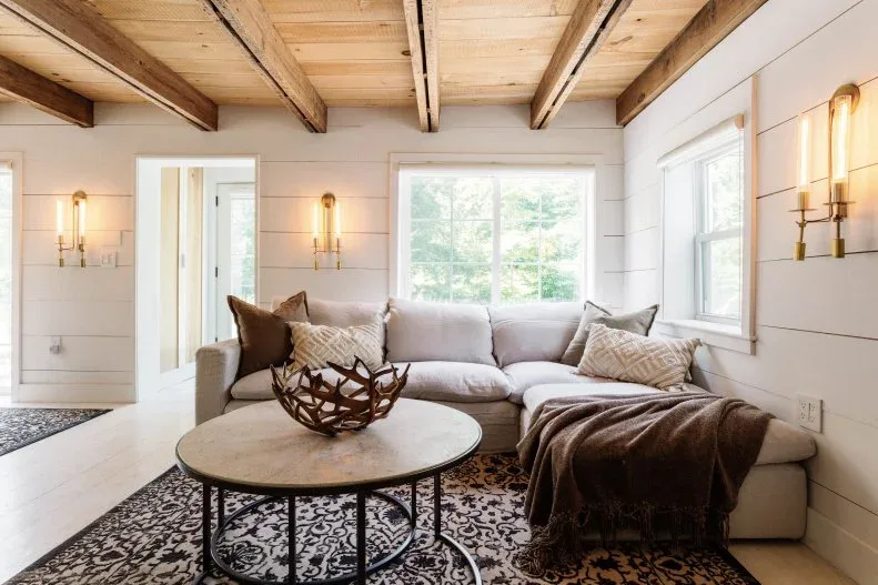 Pale sectional beneath low wood ceiling and exposed beams, with round coffee table, patterned rug and wall lights