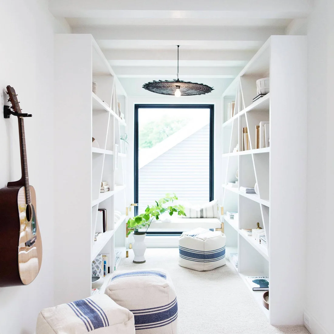 White shelves flank a tall black-framed window with a pale chair, striped poufs and a guitar on the nearby wall.