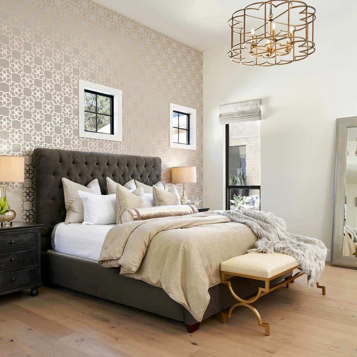 Pale looping wallpaper behind a gray tufted headboard and two small high windows with white trim.