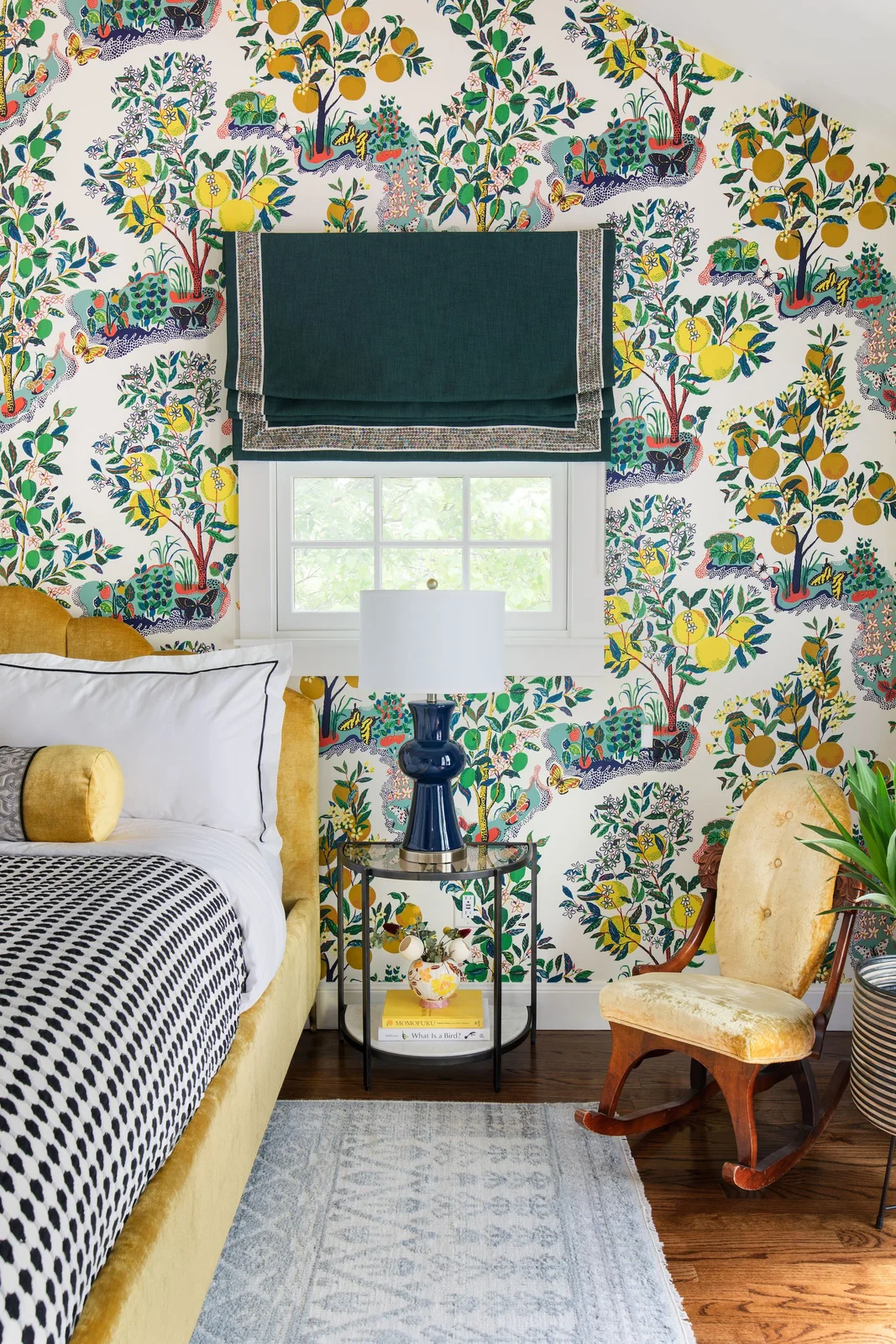 Yellow and green botanical wallpaper surrounds a yellow upholstered headboard and rocking chair with a dark green blind and dotted throw.