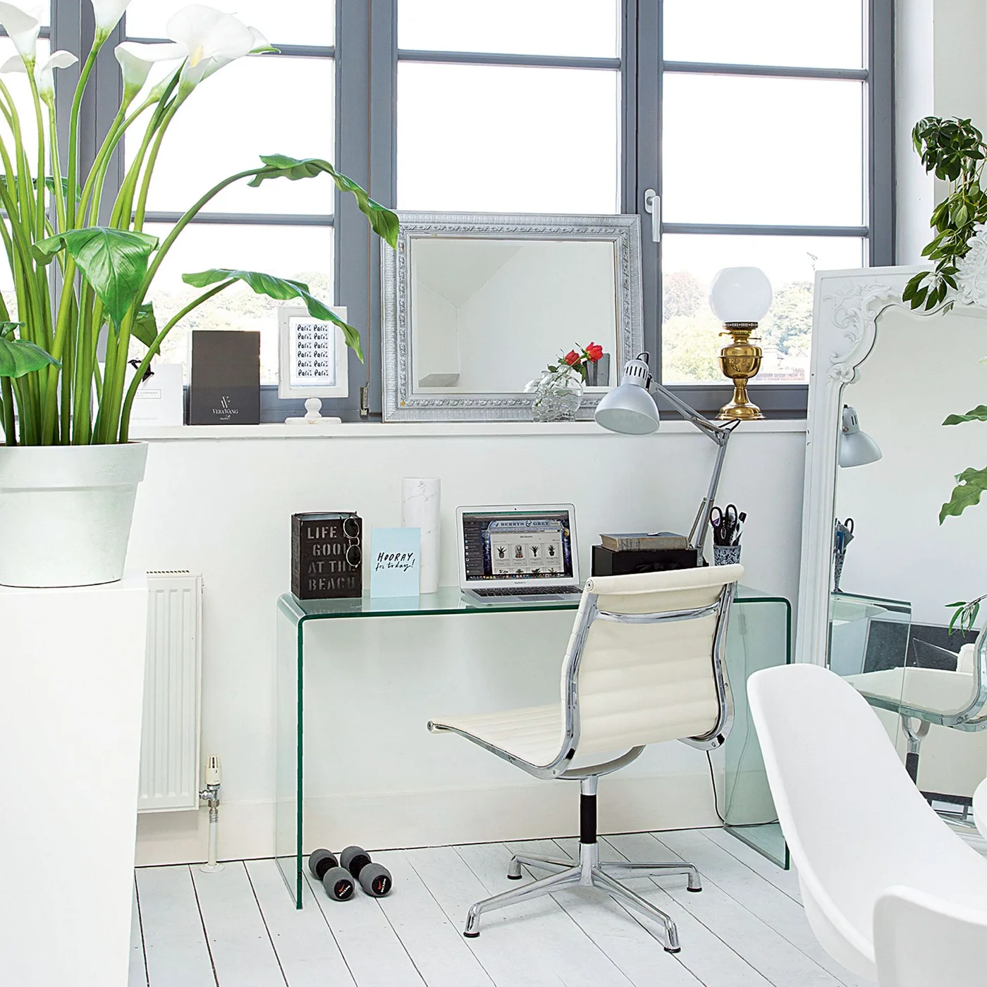 A transparent desk and white swivel chair sit below dark gray window frames beside a pale gray adjustable lamp and white painted floor