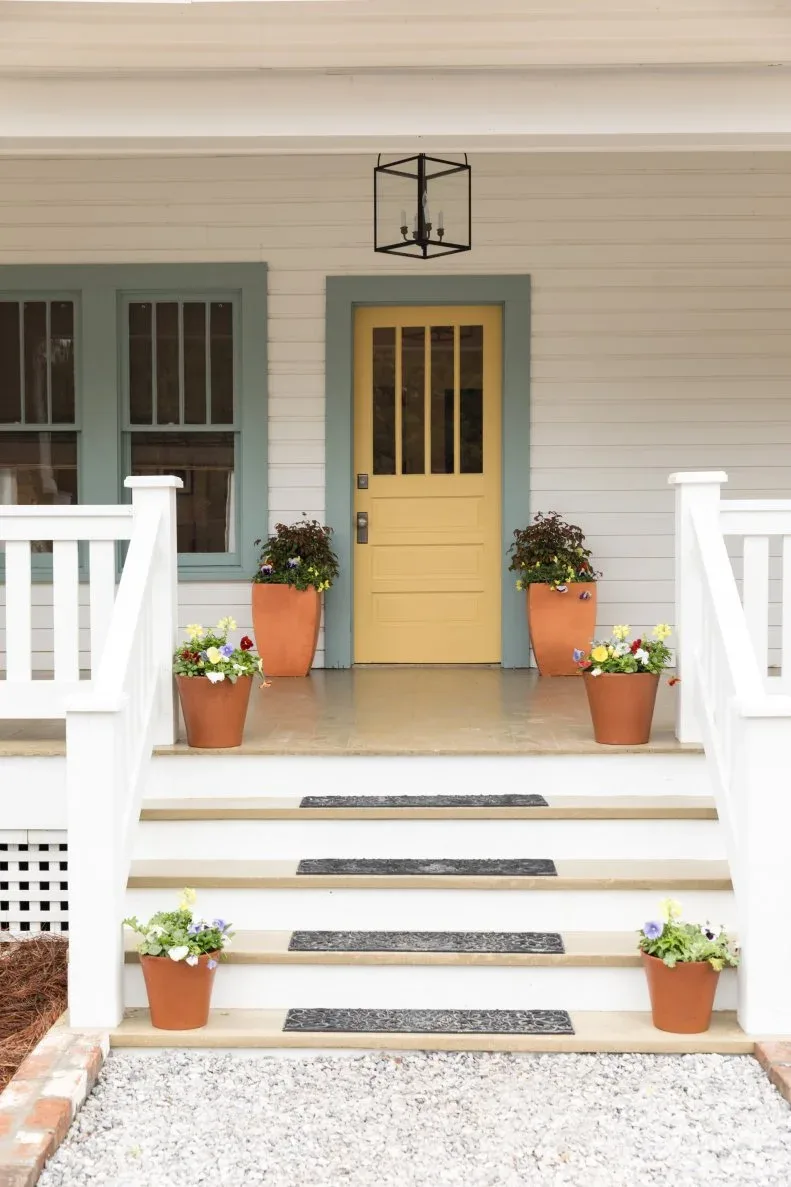 Yellow glazed front door with muted green trim, white porch rails and flowering pots