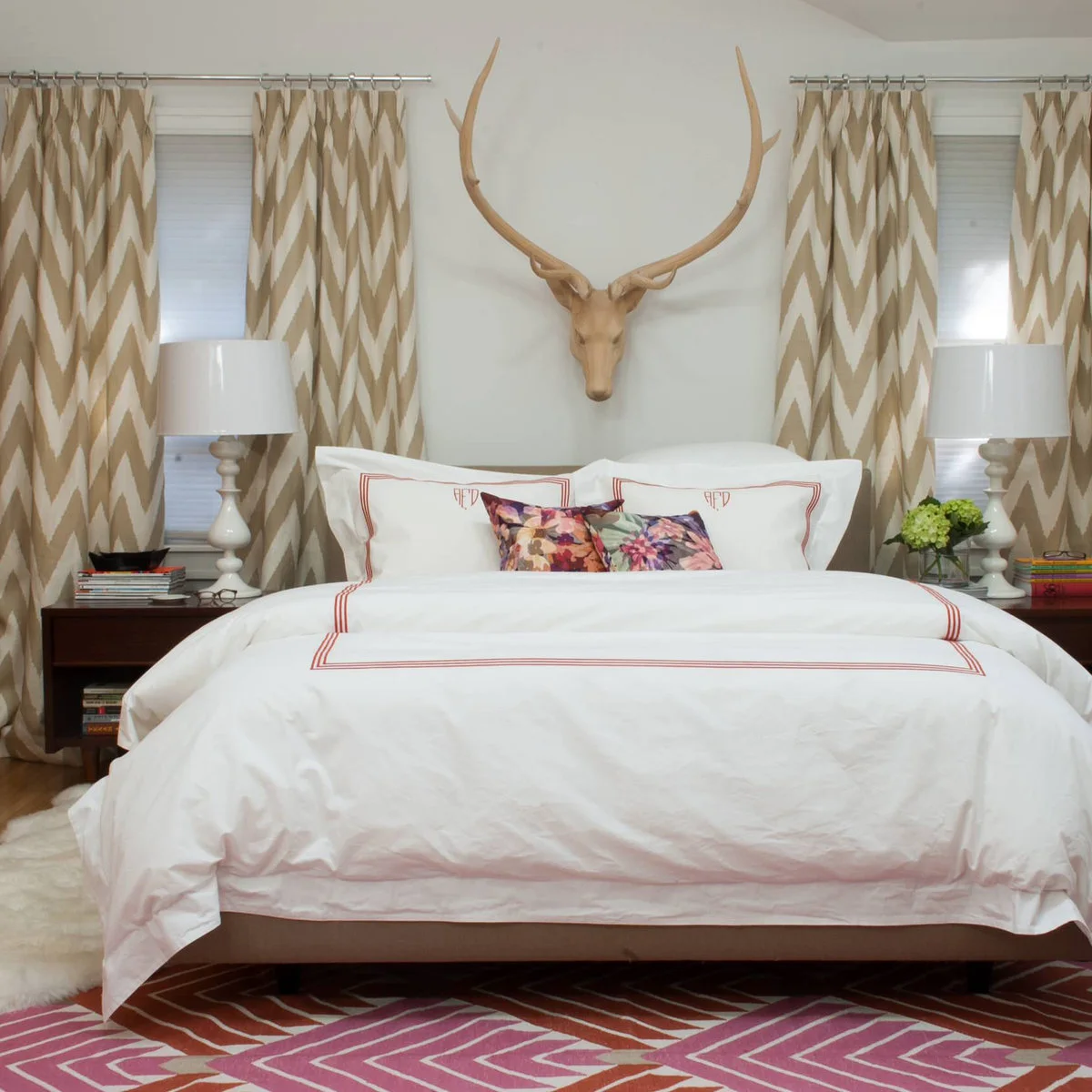 A pink, rust and cream geometric rug appears below a white bed between patterned curtains and dark bedside tables.