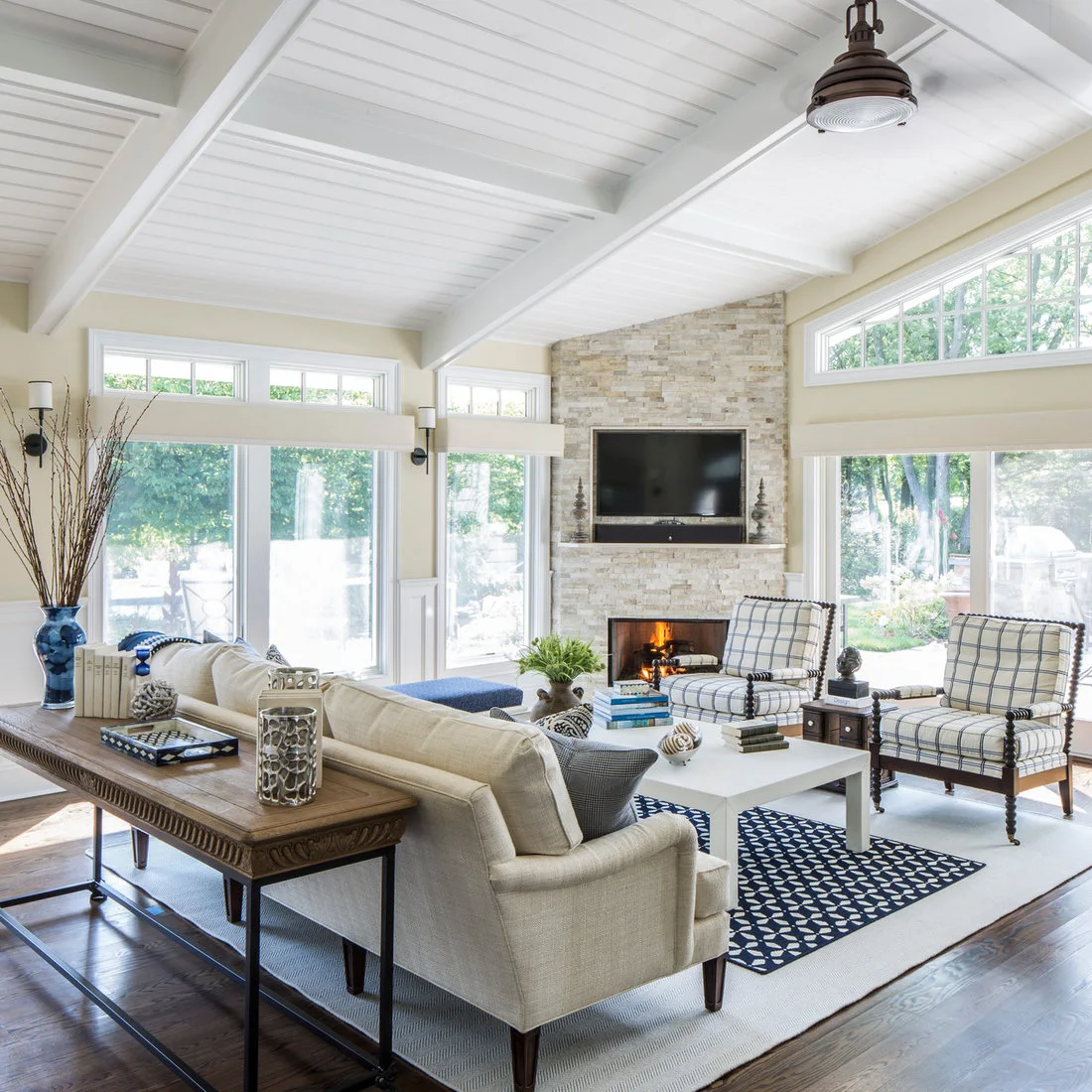 A television recessed above a pale textured fireplace in a bright room with checked chairs, a cream sofa and blue accents.