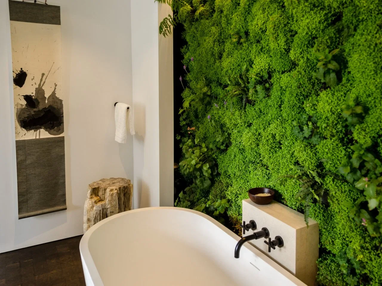 A dense green planted wall rises behind a pale tub and dark faucet beside a rough timber stool.