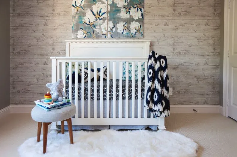 Pale paneled crib beneath floral picture against textured-looking wall with darker staged throw and pale round stool.