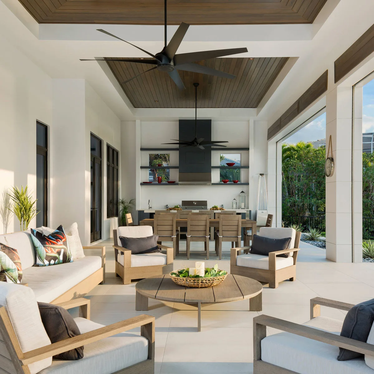 Long covered outdoor room with wood-framed lounge furniture, dining table and kitchen at the far end