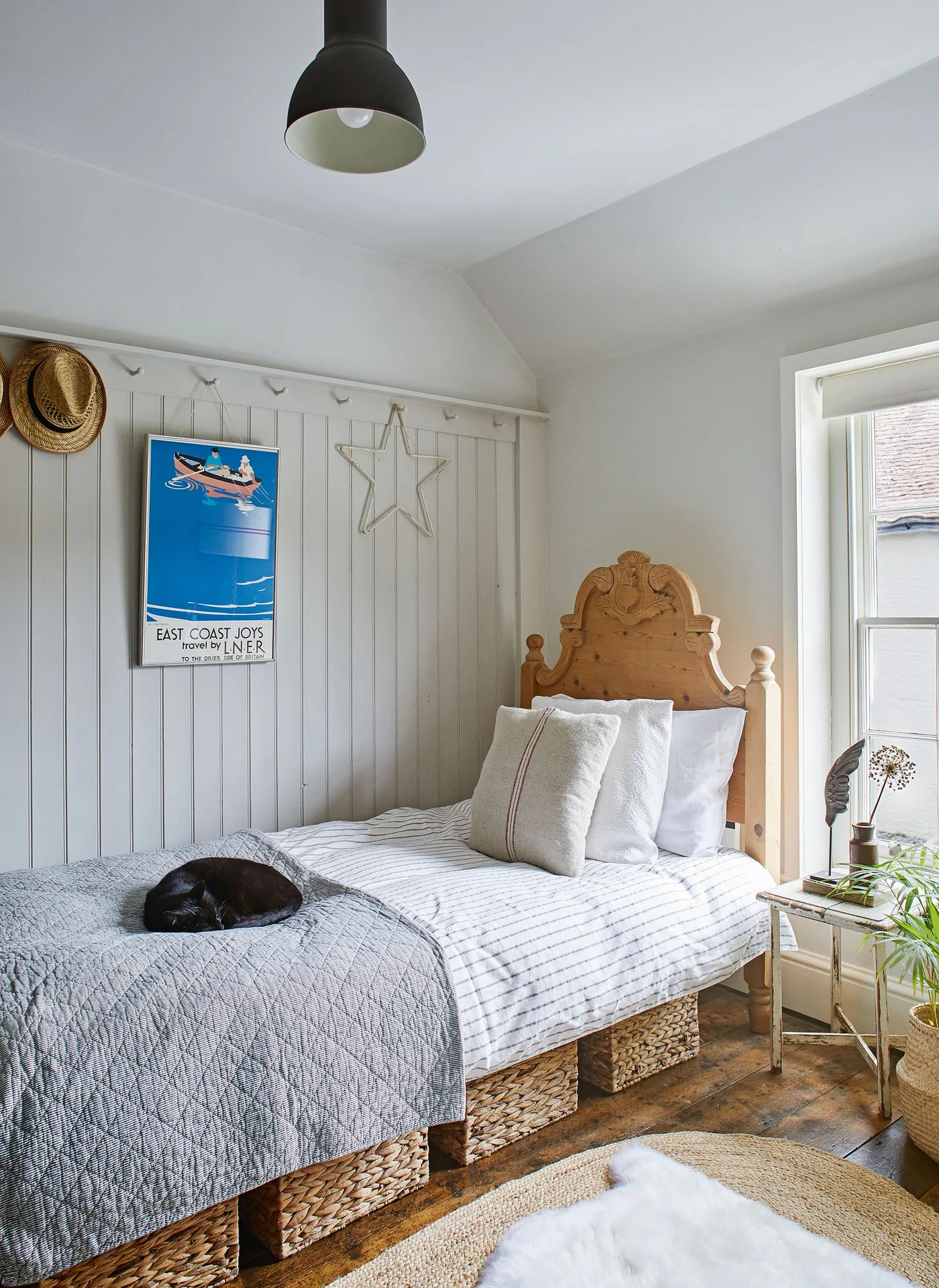 Small country bedroom with a carved wooden single bed, striped pale bedding, woven baskets underneath, pale wall boards and a blue framed print.