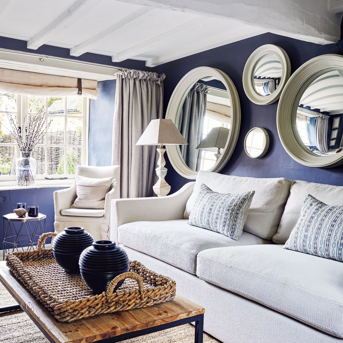 Different pale-framed mirrors hang against a dark blue wall above a white sofa beside a window and white ceiling beams