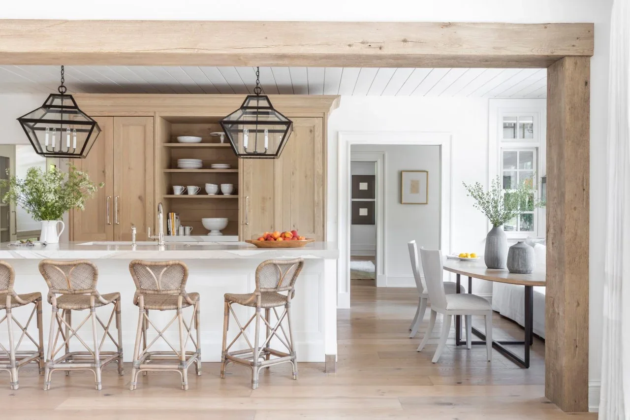 Pale wood cupboards and open dish shelves form a dresser-like wall behind a white island and woven stools.