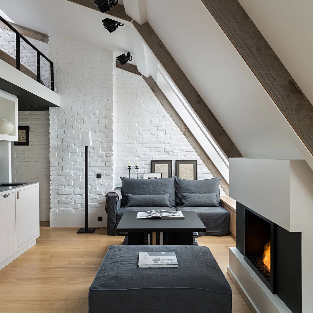 A dark gray sofa and ottoman beneath sloping beams beside white brick and a black edged fireplace