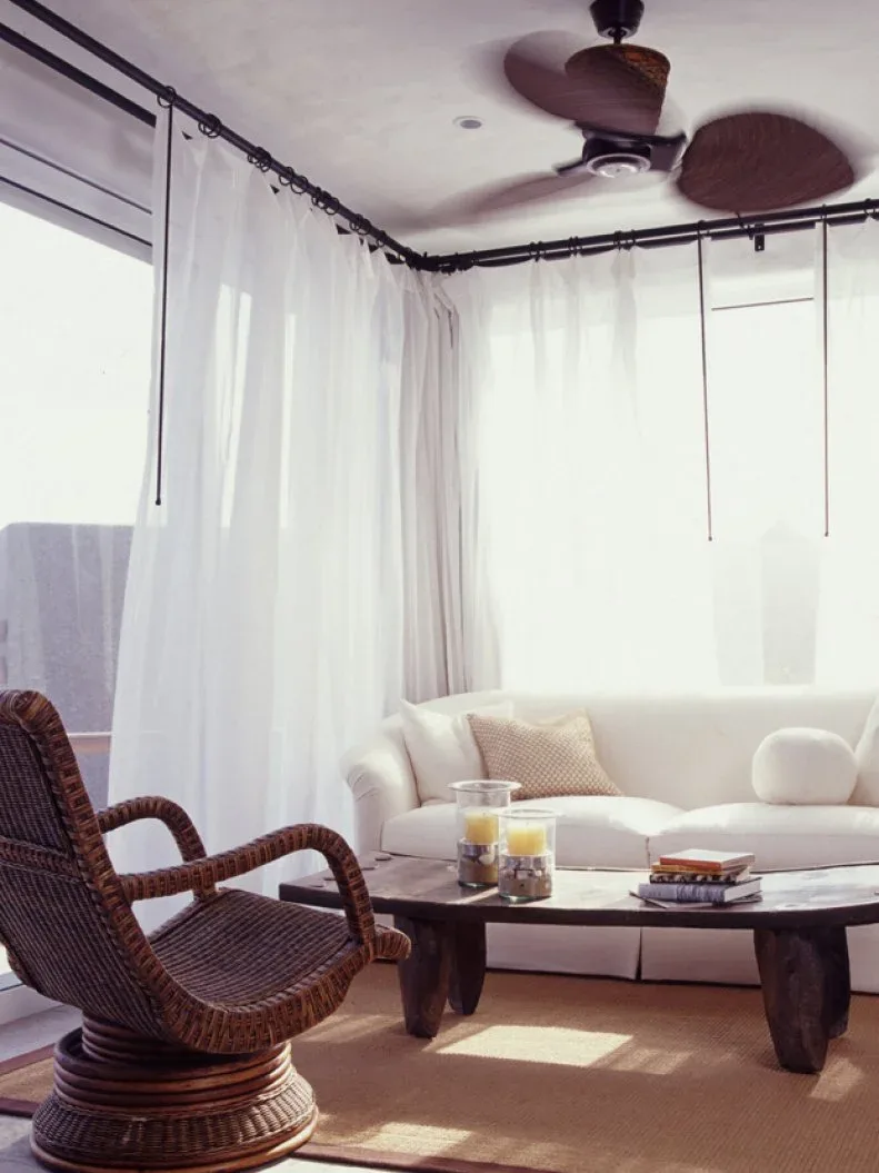 Pale sofa and dark woven chair around a low wooden table with sheer curtains and a dark ceiling fan.