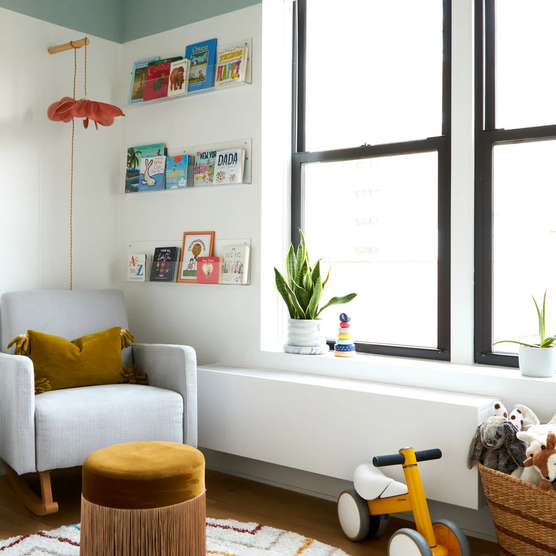 A gray nursery chair with a mustard cushion and round pouf stands beside clear book ledges, a large black-framed window and plants.