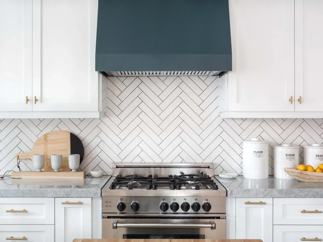 White herringbone kitchen tiles with dark joints sit behind a range beneath a blue-gray hood.