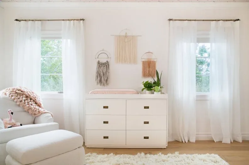 Pale dresser with three fringed wall hangings between windows beside pale chair and ottoman on wooden floor.