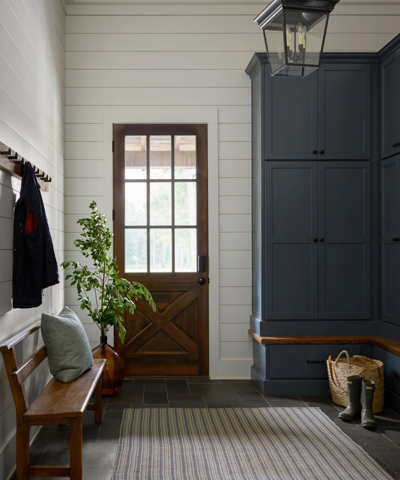 Dark blue-gray corner cupboards and a low drawer stand opposite a wood bench in a white-paneled mudroom with a glazed wood door.
